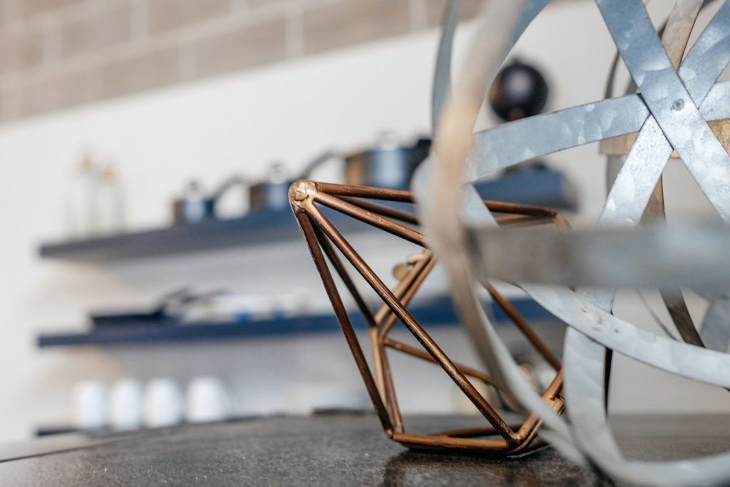 Primer plano de una estructura de alambre de cobre, posiblemente una obra de arte o un mueble, en una habitación con una rueda metálica y estanterías al fondo.