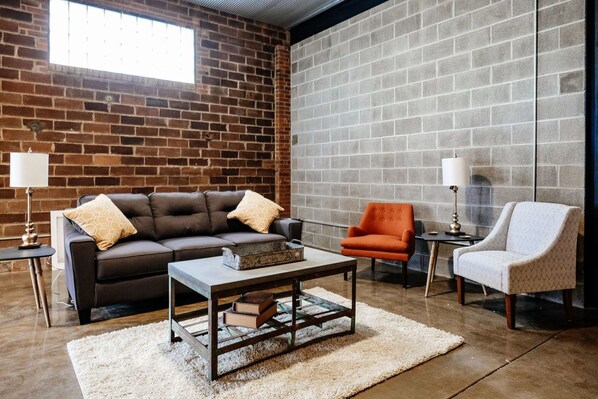 A living room with a brick wall, a grey sofa with cushions, an orange armchair, a white armchair, and a coffee table with books.