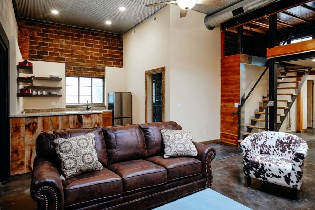 A spacious living area with a brown leather sofa, a patterned armchair, and a kitchen area with a refrigerator and shelves.
