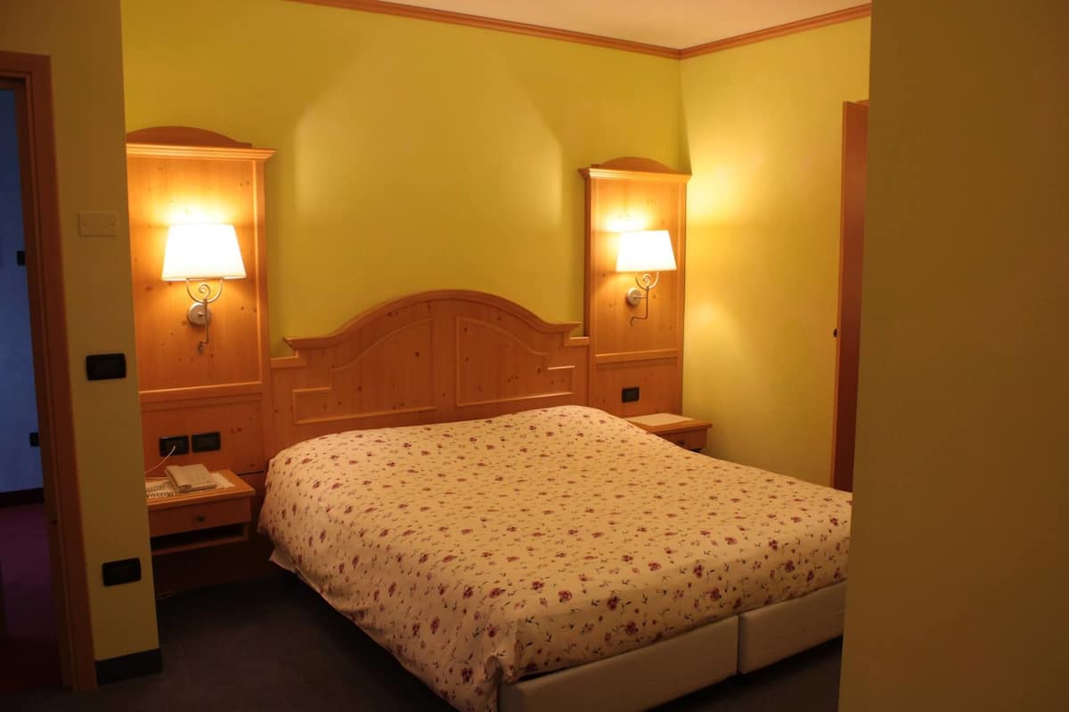 Ein Hotelzimmer mit hölzernem Kopfteil, floralem Bettbezug, zwei Nachttischlampen, einem Nachttisch mit Telefon und einer Tür links.