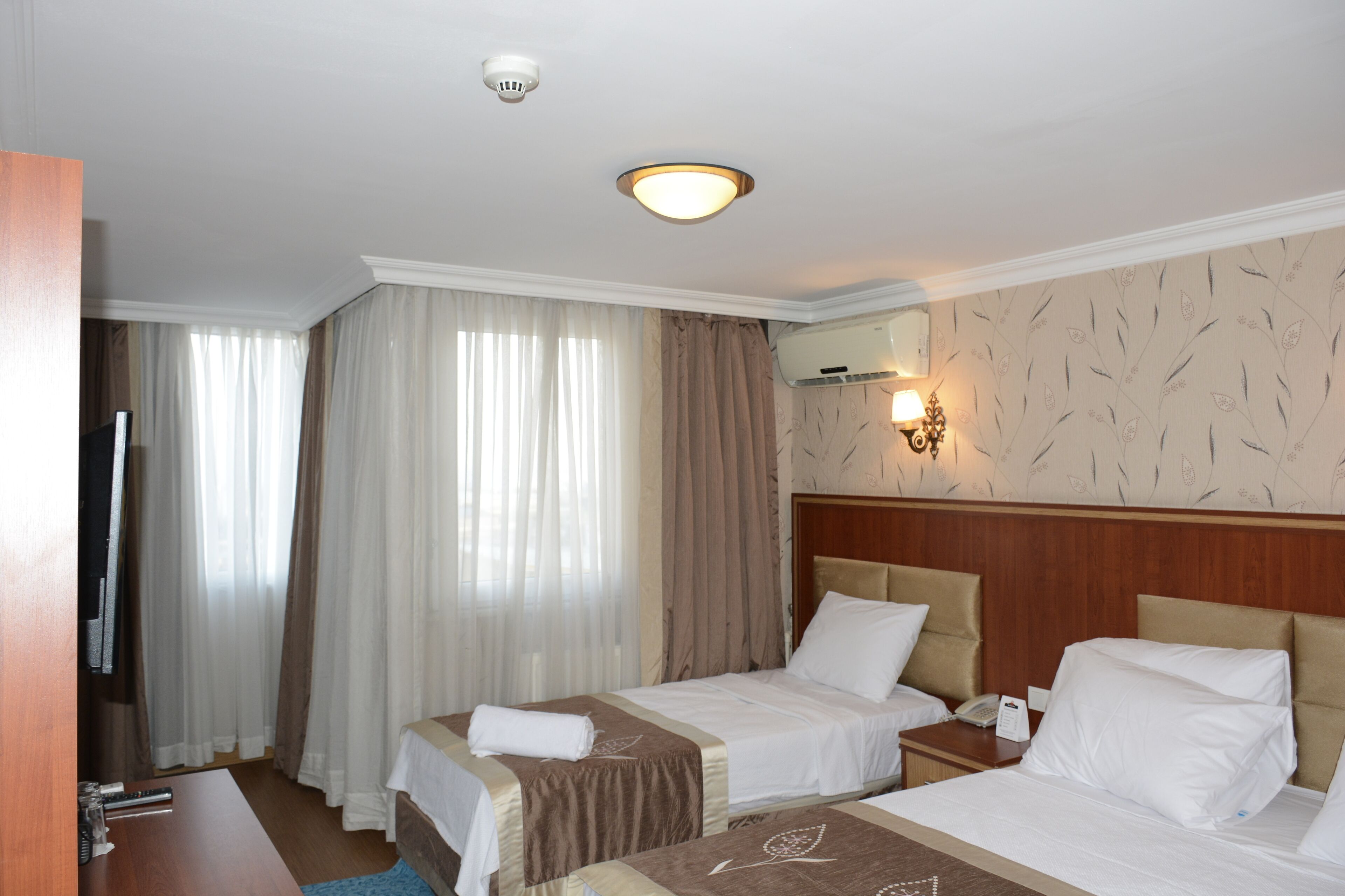 Hotellihuone, jossa on kaksi sänkyä, televisio, yöpöytä, lamppu ja ikkuna, jossa on verhot.