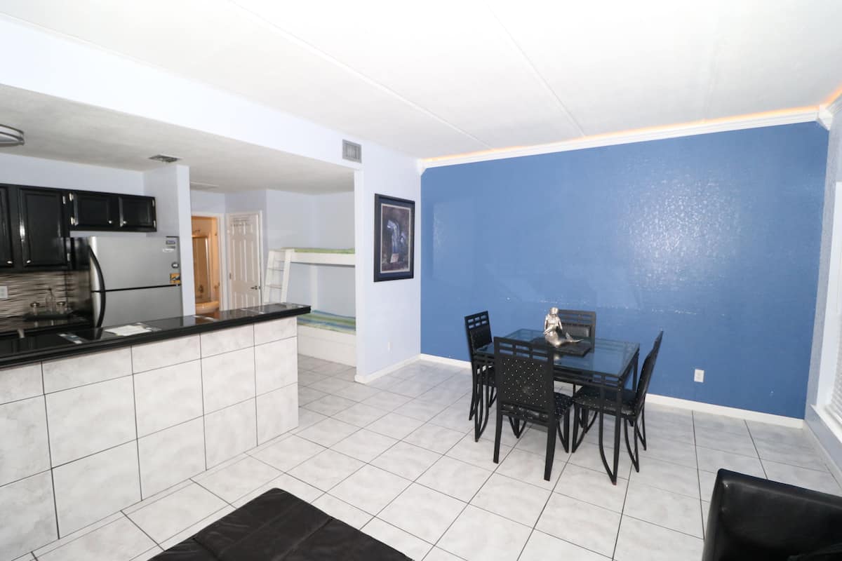 Cocina moderna con armarios negros y un refrigerador de acero inoxidable. Hay una zona de comedor con una mesa de vidrio y sillas negras. Las paredes están pintadas de azul y el suelo está cubierto de baldosas.