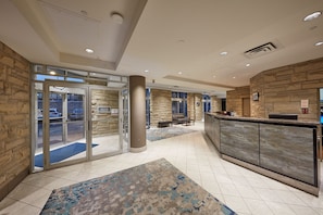 A lobby with a reception desk, glass doors leading outside, and a stone wall. - The Falls Hotel & Inn (Niagara Falls)