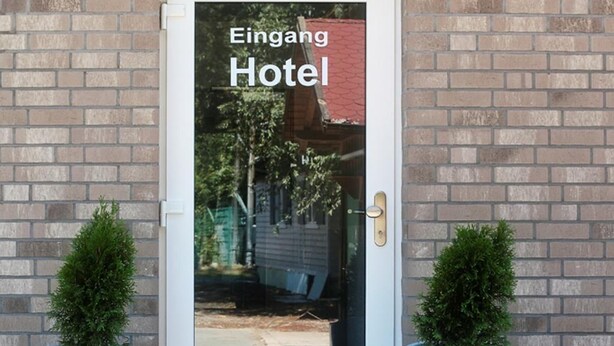 Entrada de hotel con puerta de vidrio, dos plantas en macetas y pared de ladrillo.