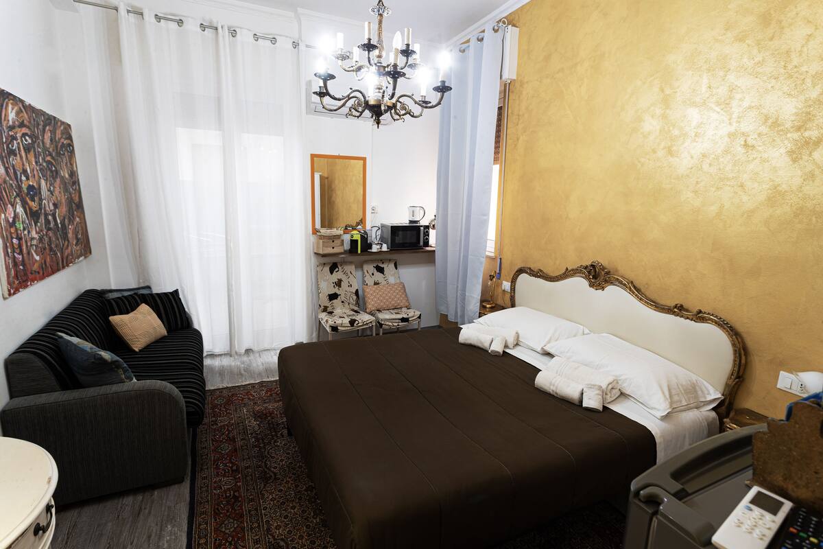 A hotel room with a large bed, a sofa, a desk with a computer, and a chandelier.