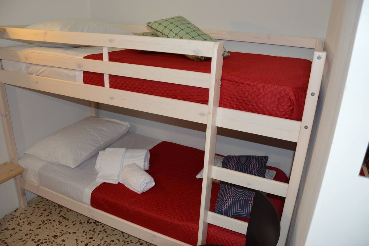 A bunk bed with red bedding and pillows, a small shelf, and a wooden floor.