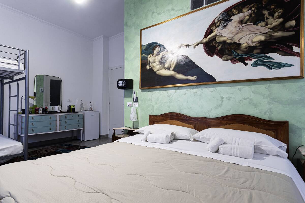 A hotel room with a bunk bed, a large bed with white linens, a painting of angels on the wall, and a small desk with a lamp.