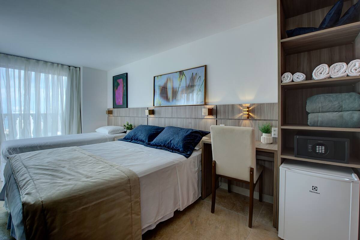 Ein Hotelzimmer mit zwei Betten, einem Schreibtisch, einem Stuhl, einem Kühlschrank und einem Regal mit Handtüchern.