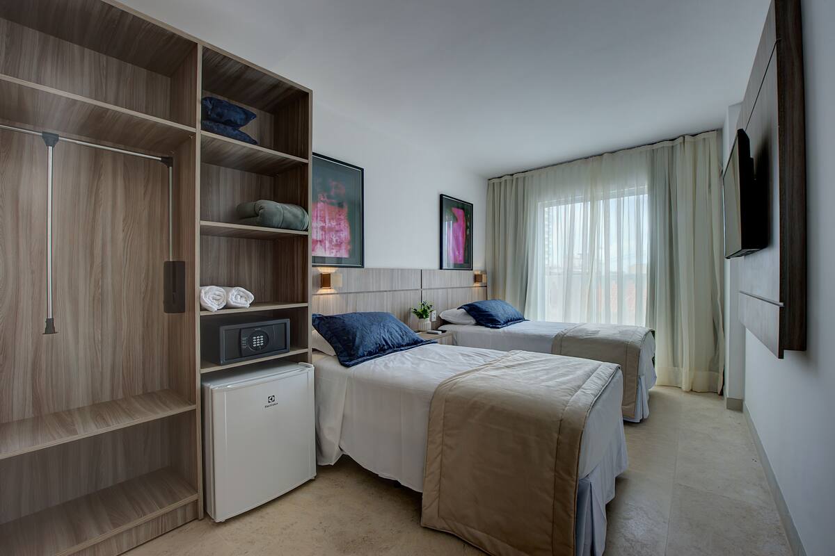 Ein Hotelzimmer mit zwei Betten, einem Schrank, einem kleinen Kühlschrank und einem an der Wand befestigten Fernseher.