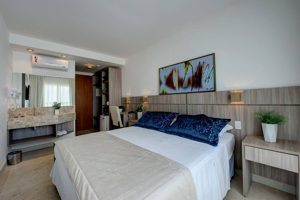 Ein modernes Hotelzimmer mit einem großen Bett, einem fest installierten Schreibtisch, einem Schminktisch mit Spiegel und einer wandmontierten Klimaanlage.