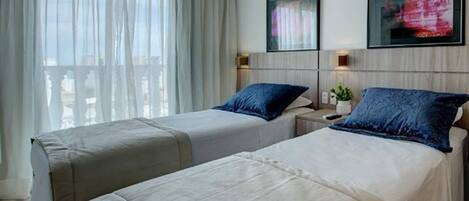 A hotel room with two beds, each with a pillow and a bedside table. There is a window with sheer curtains, a framed picture on the wall, and a small plant on the bedside table.