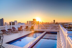 Una piscina en la azotea con sillones de descanso y vistas a un paisaje urbano al atardecer.