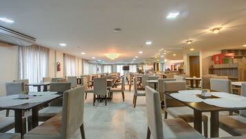 Een ruime hotel eetzaal met meerdere tafels en stoelen, een kroonluchter en een Coca-Cola koelkast.