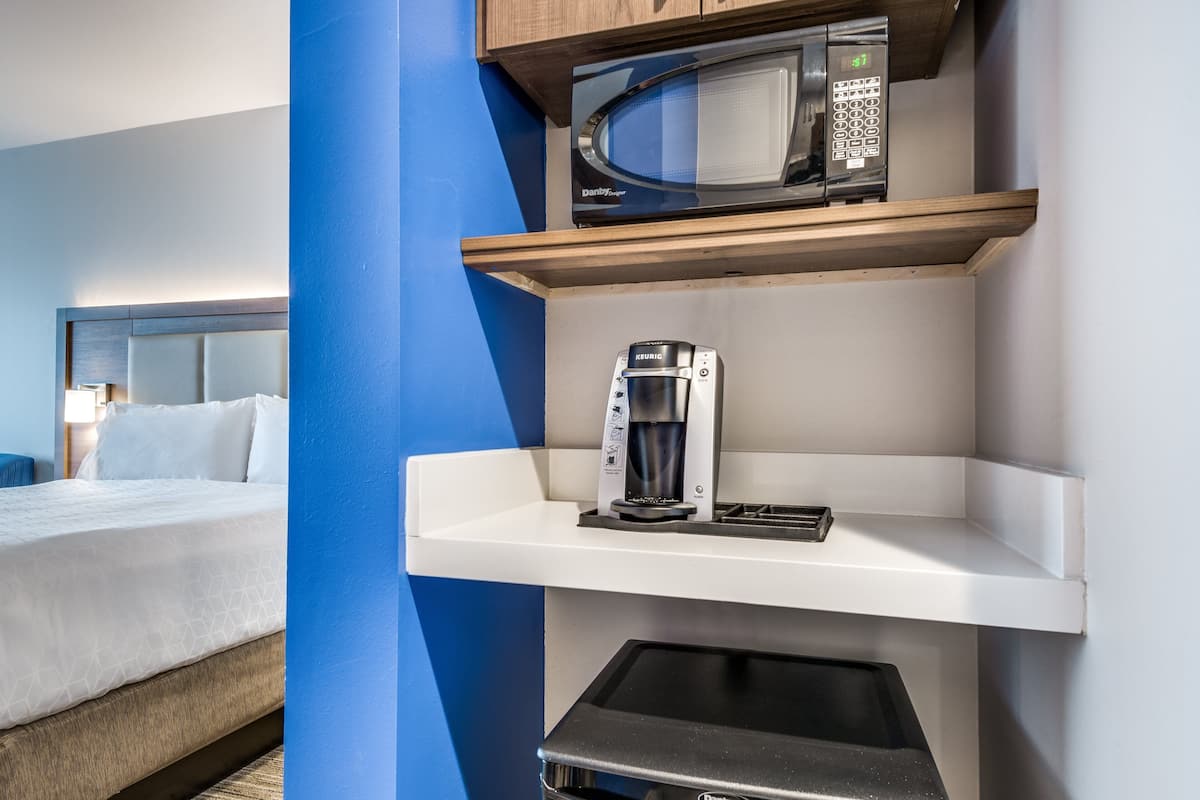 A hotel room with a bed, a microwave, a coffee maker, and a small shelf.