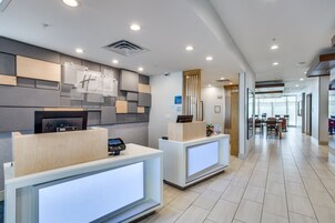 A modern hotel reception area with two reception desks, a digital check-in station, and a view into the dining area.