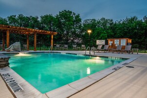 A swimming pool with lounge chairs and a covered area, surrounded by trees.