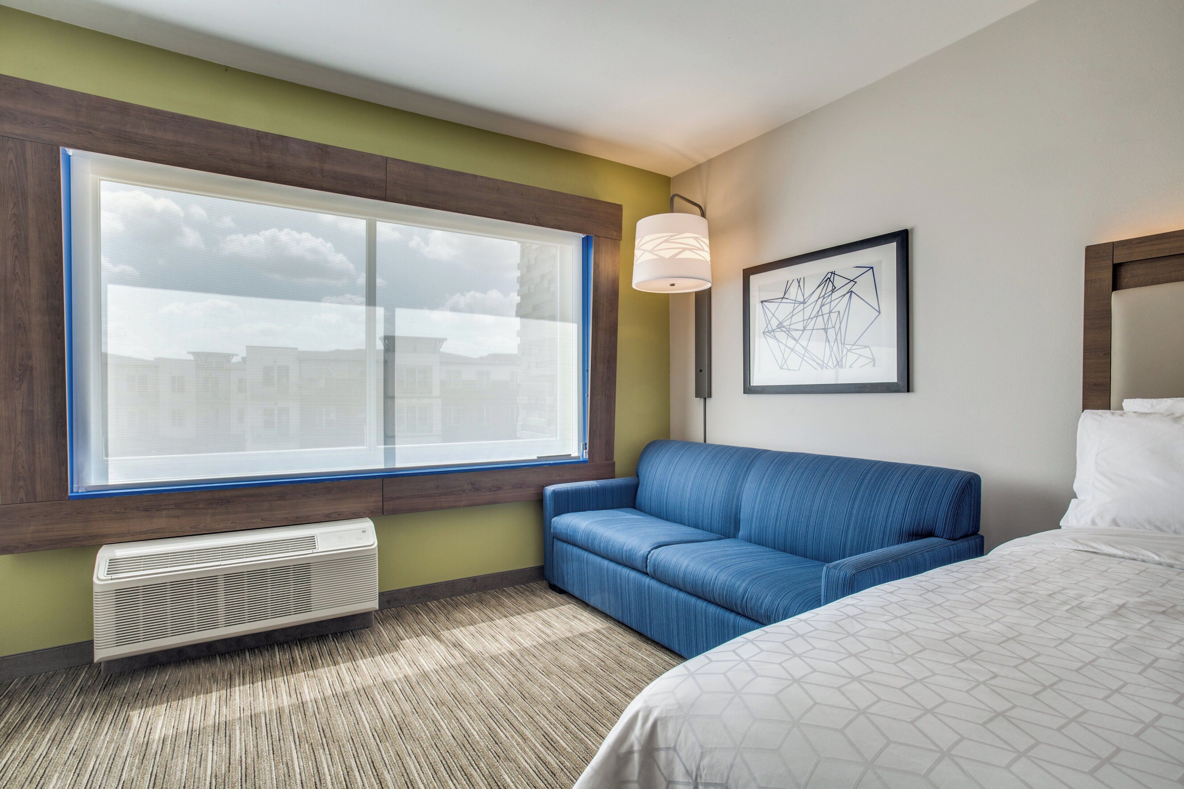 Habitación de hotel con un ventanal grande, un sofá azul, una cama con colcha blanca estampada, una lámpara de pared y una obra de arte abstracta enmarcada.