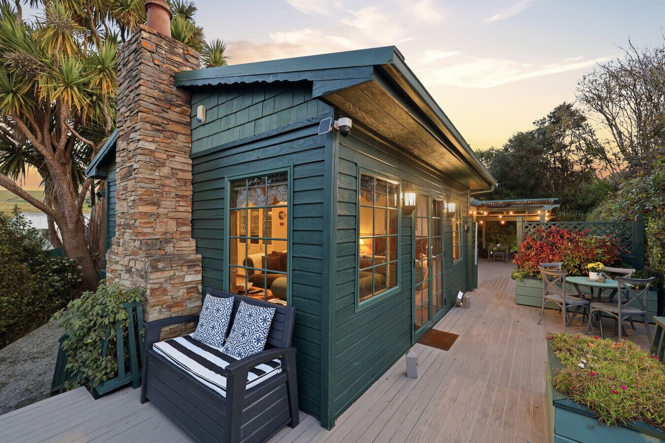 A cozy cabin with a stone chimney, a deck with outdoor furniture, and a lush garden.