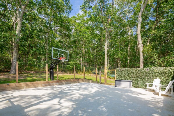 An outdoor basketball court with a wooden fence, a basketball hoop, and a hedge in the background.