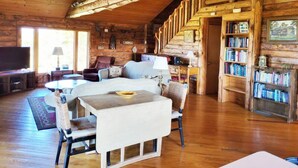Interior acogedor de una cabaña de troncos, con una escalera de madera, un televisor, una zona de comedor y una estantería.