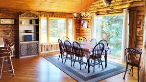 Interior de una cabaña de madera con una mesa de comedor, sillas, una estantería y un ventanal grande.