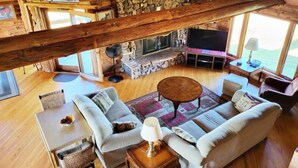 Un salón amplio con chimenea de piedra, vigas de madera, un televisor grande y asientos cómodos.