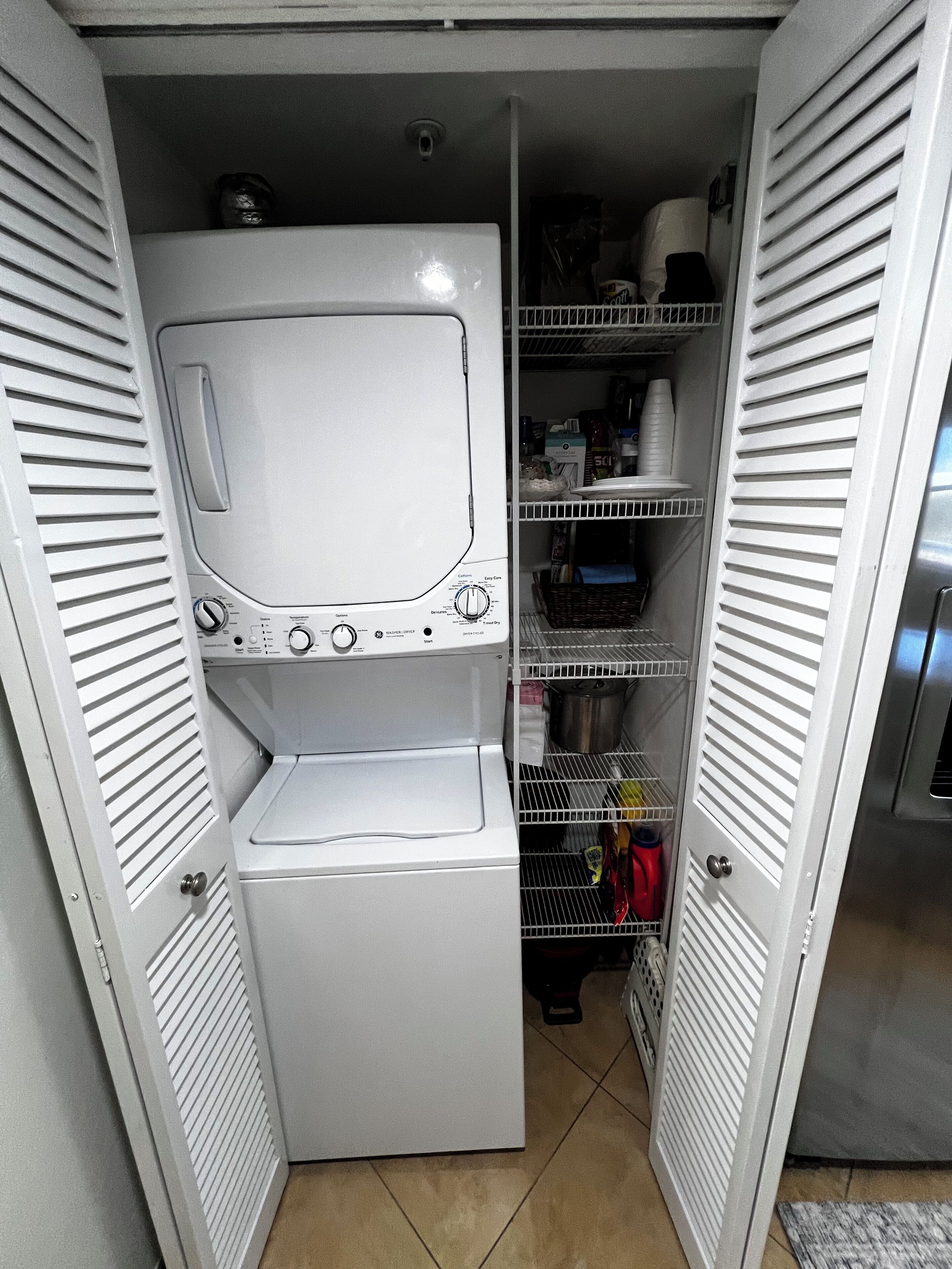 A compact laundry room with a front-loading washer and dryer, metal shelving with various items, and a tiled floor.