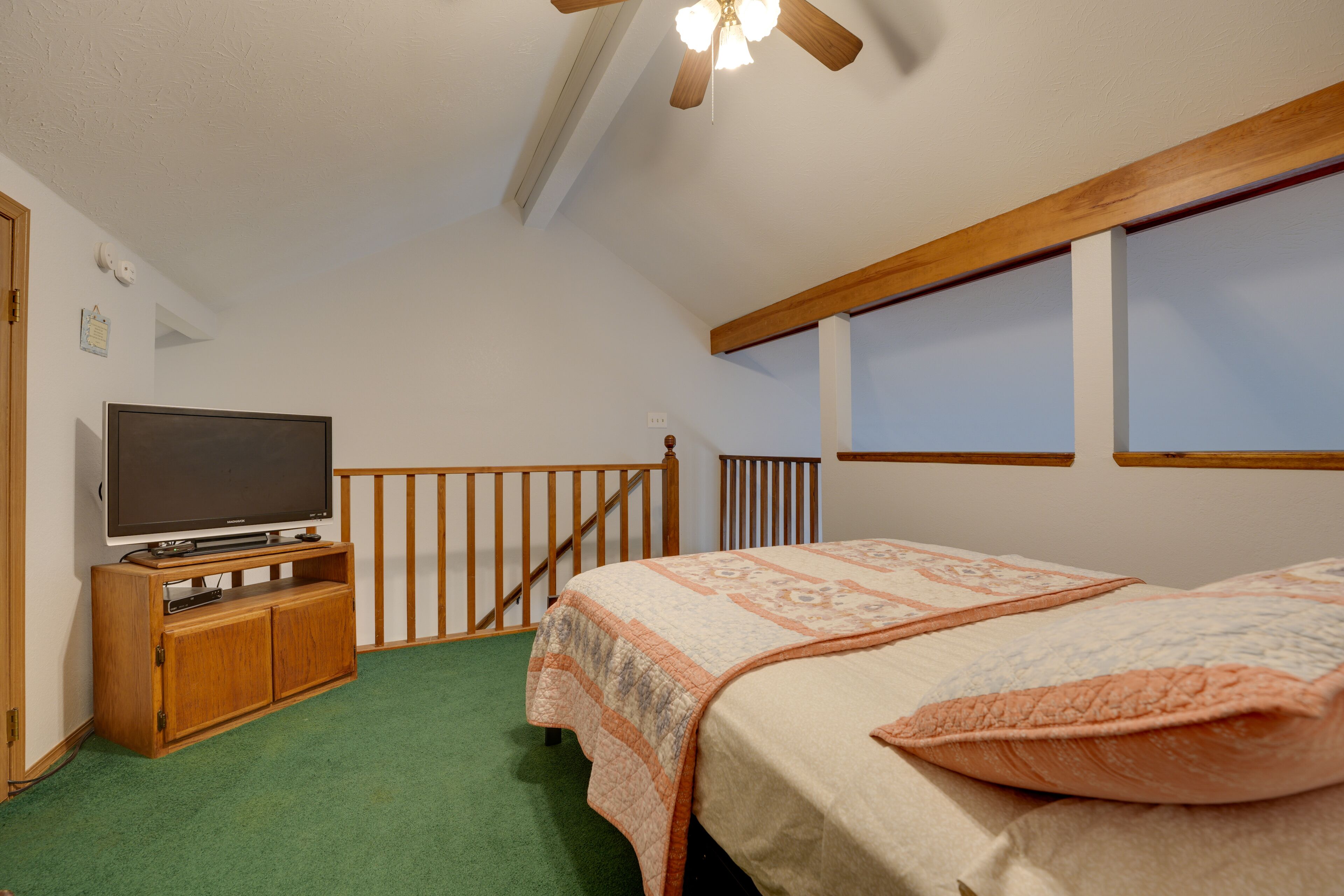Une chambre à coucher avec un lit, un téléviseur posé sur un meuble en bois, une fenêtre et un ventilateur de plafond.