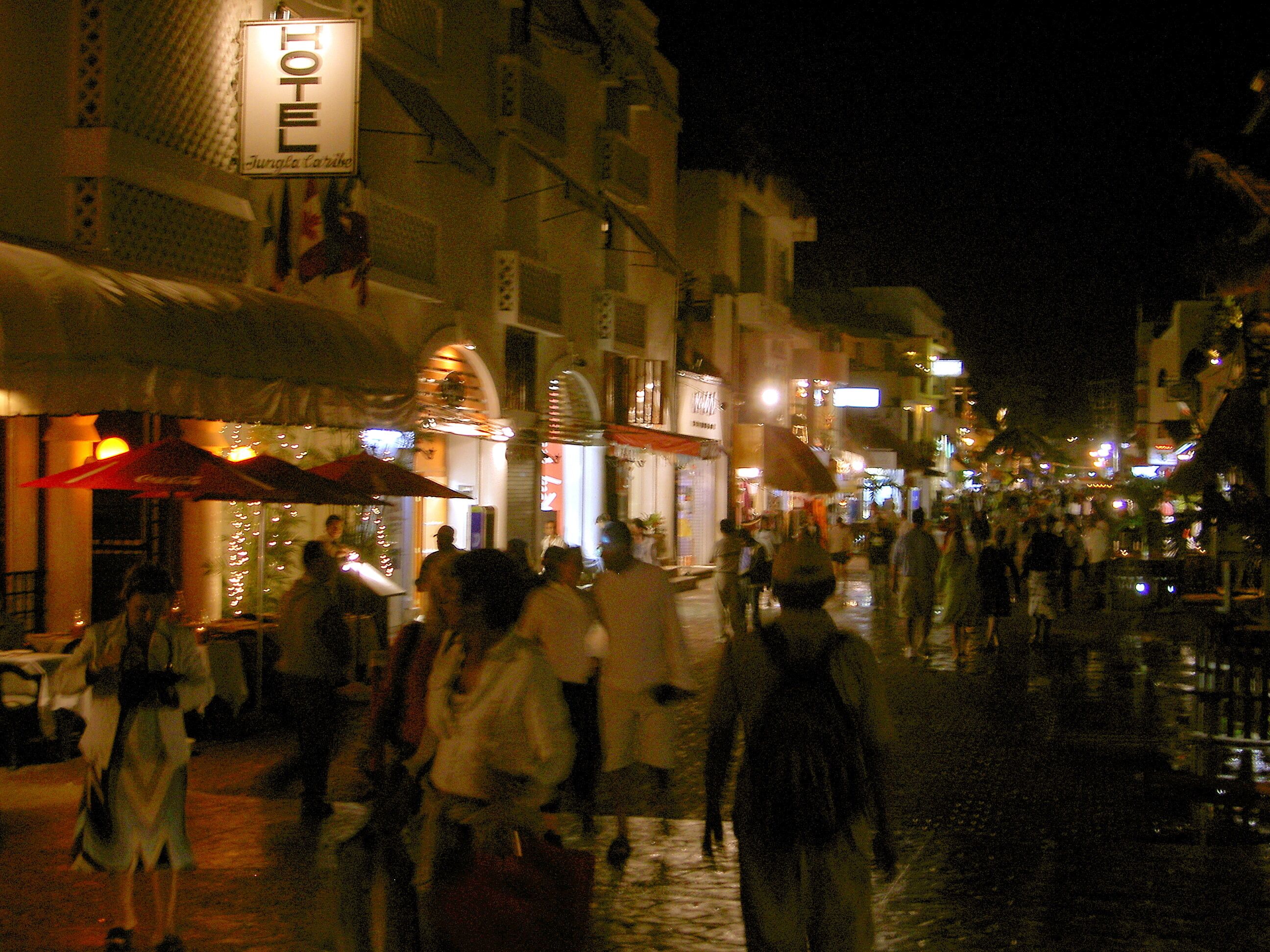 Une rue animée la nuit, avec des enseignes lumineuses et des tables installées à l’extérieur.
