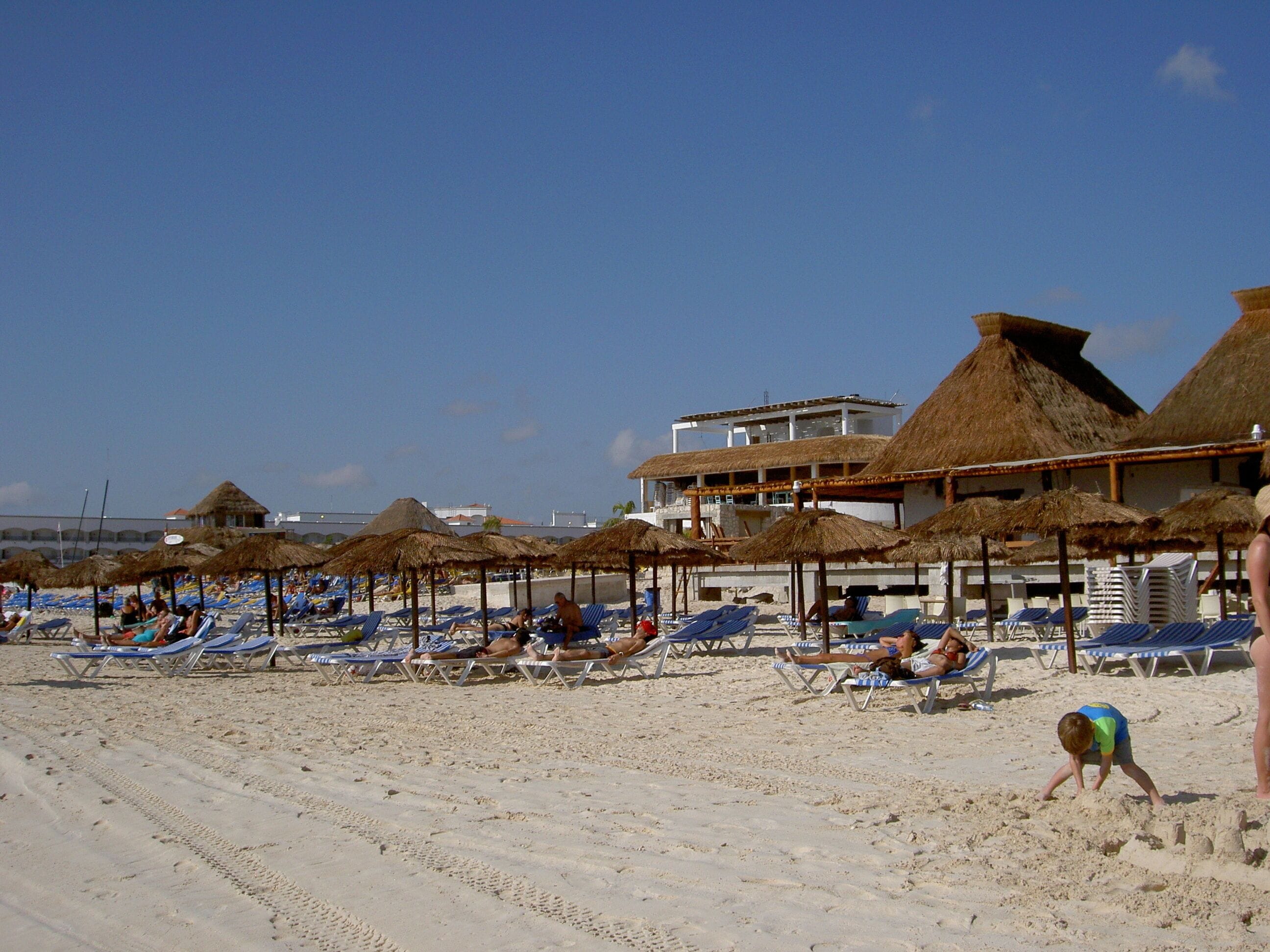 A beach with sun loungers and thatched umbrellas, a building with a thatched roof, and a clear sky.