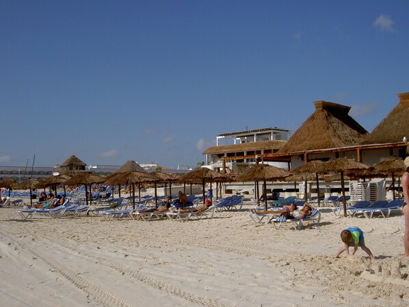 A beach with sun loungers and thatched umbrellas, a building with a thatched roof, and a clear sky. - Steps from 5th and Mamitas Beach! And the Reviews Say..... Great Location! (Playa del carmen)