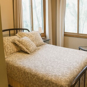 Un lit recouvert d’une couvre-lit à motifs, une table de chevet avec une lampe, et une vue sur les arbres par la fenêtre.