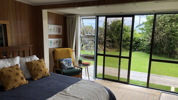 A bedroom with a large bed, a chair with a yellow blanket, a small table with a plant, and a view of a green garden through a sliding glass door.