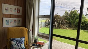 A room with a large window, a wicker chair with a cushion, a small table with a plant and a book, and framed pictures on the wall.