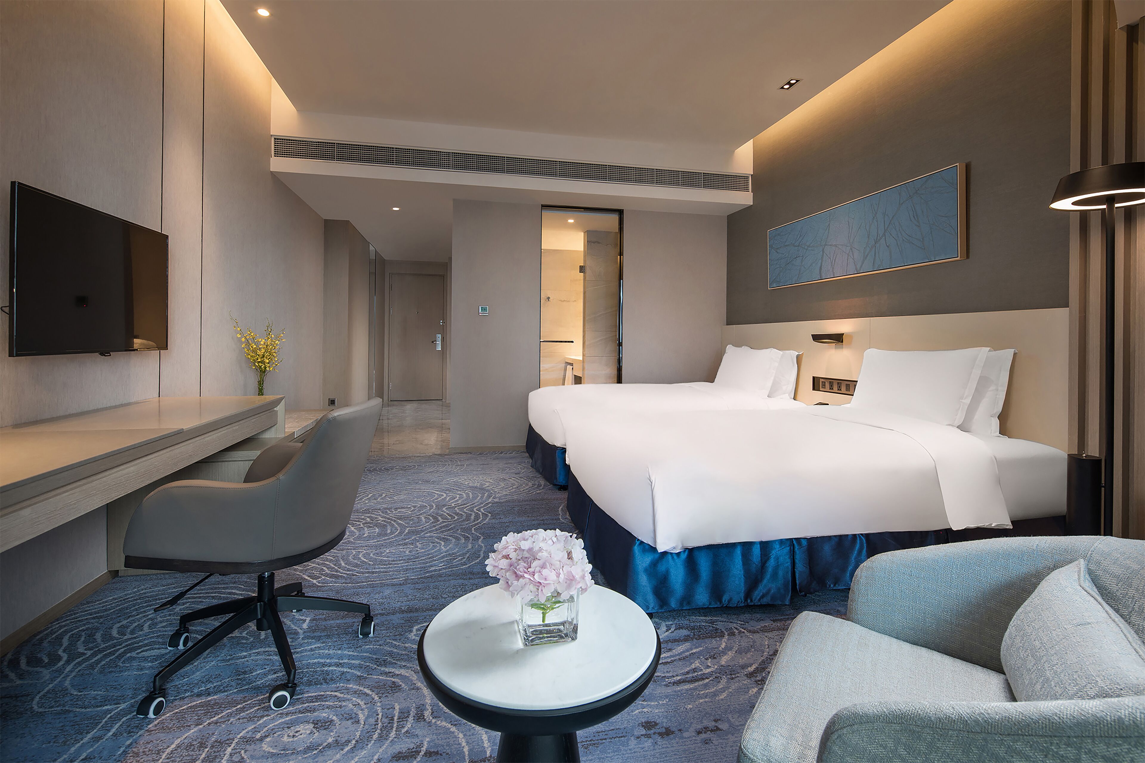 A hotel room with a bed, a desk, a chair, a small table with a vase, and a TV on the wall.