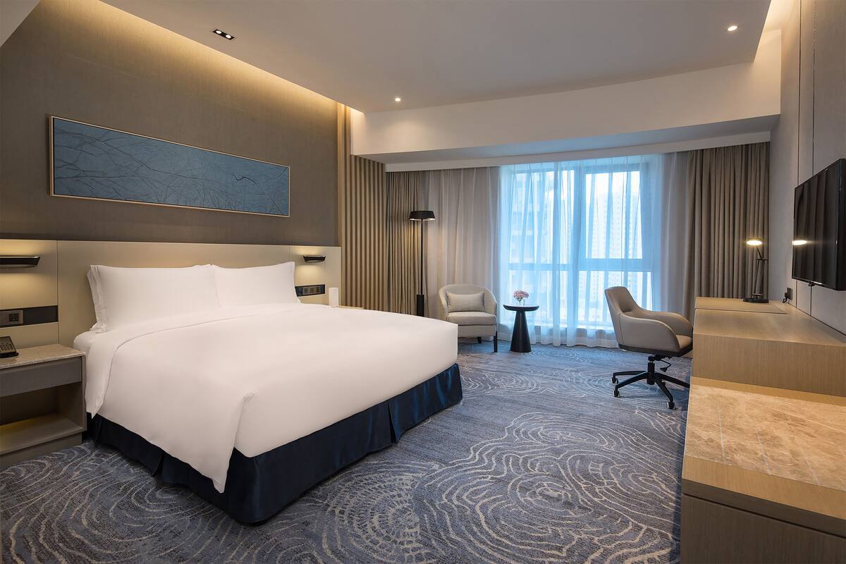 A modern hotel room with a large bed, two armchairs, a desk, and a TV.
