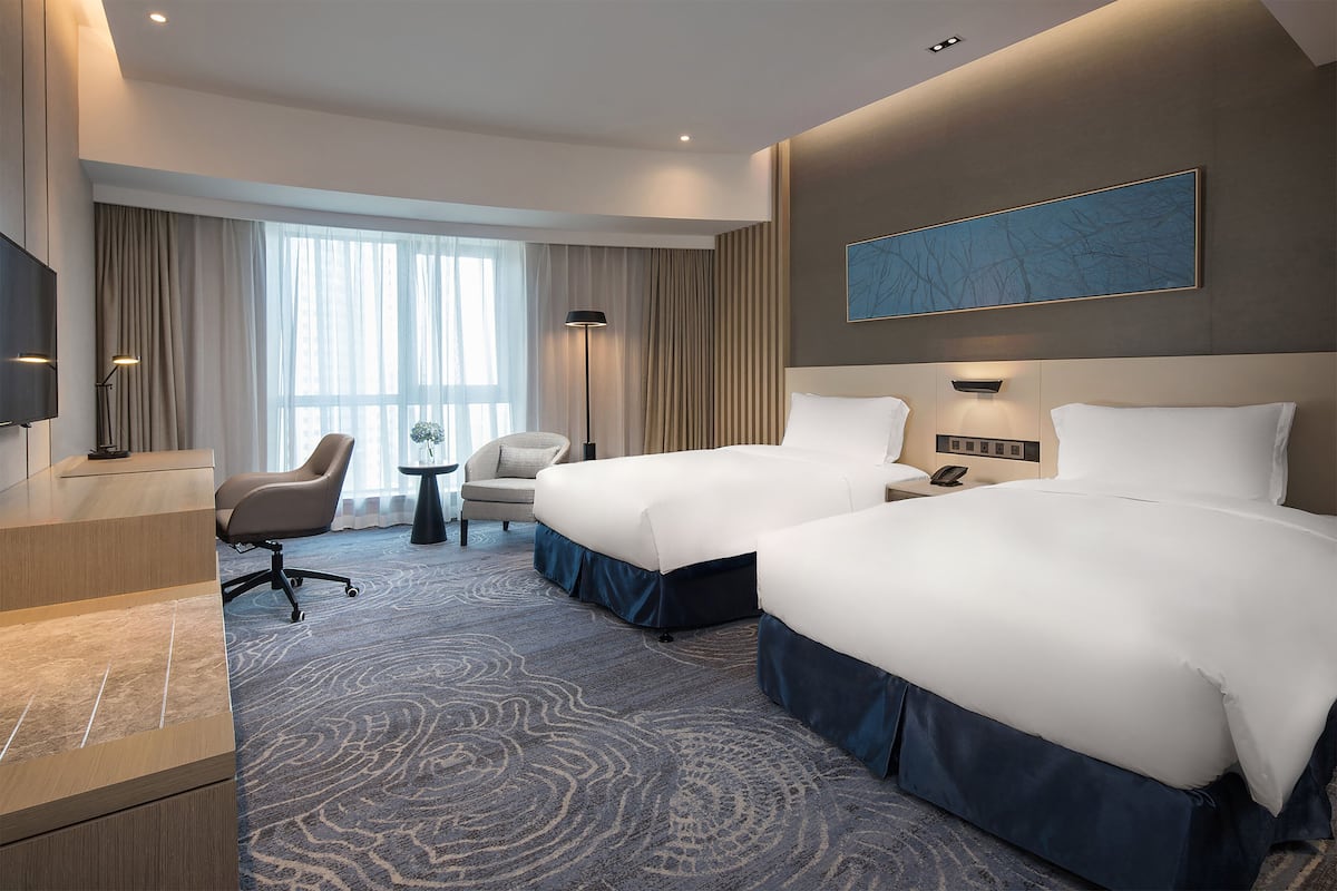 A hotel room with two beds, a desk, a chair, a lamp, and a large window with curtains.