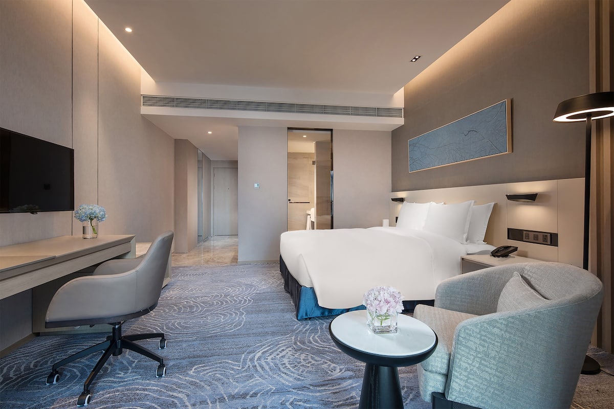 A modern hotel room with a large bed, a desk, a chair, a small table with a vase, and a flat-screen TV.