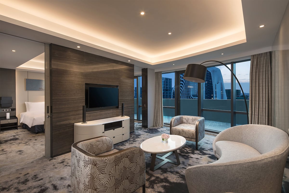 A modern hotel room with a large TV, comfortable seating, a balcony with a city view, and a large window.