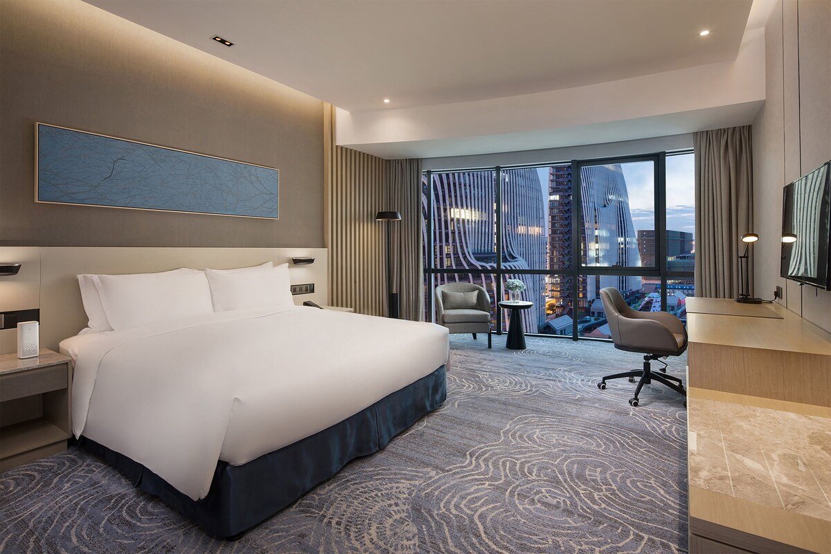 A hotel room with a large bed, a desk, a chair, a TV, and a view of the city through large windows.