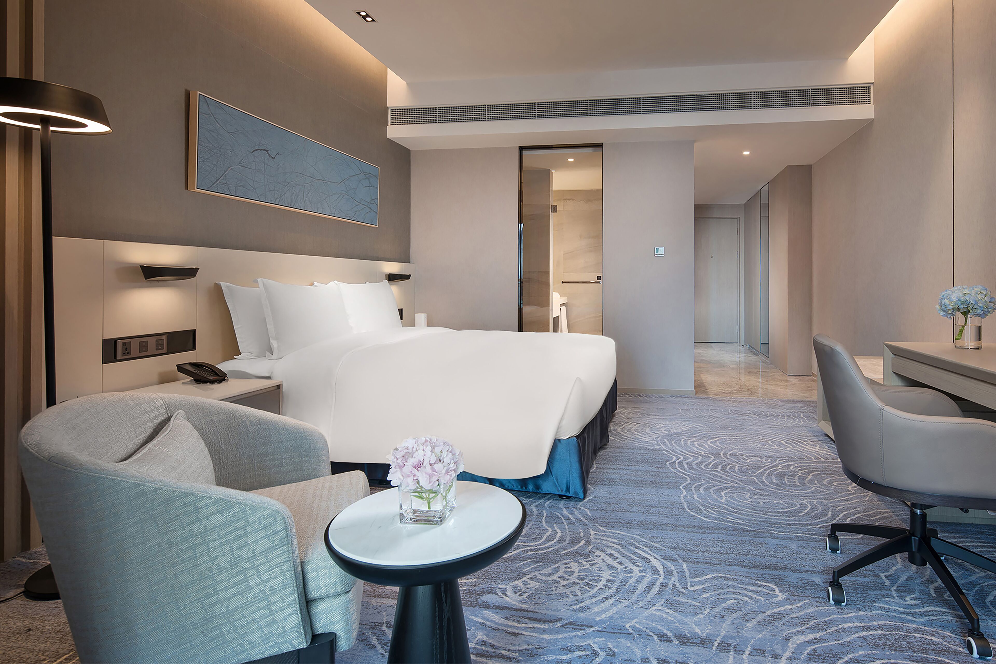 Ein modernes Hotelzimmer mit einem großen Bett, einem Schreibtisch, einem Sessel, einem kleinen Tisch mit Blumenvase und einem Gemälde an der Wand.