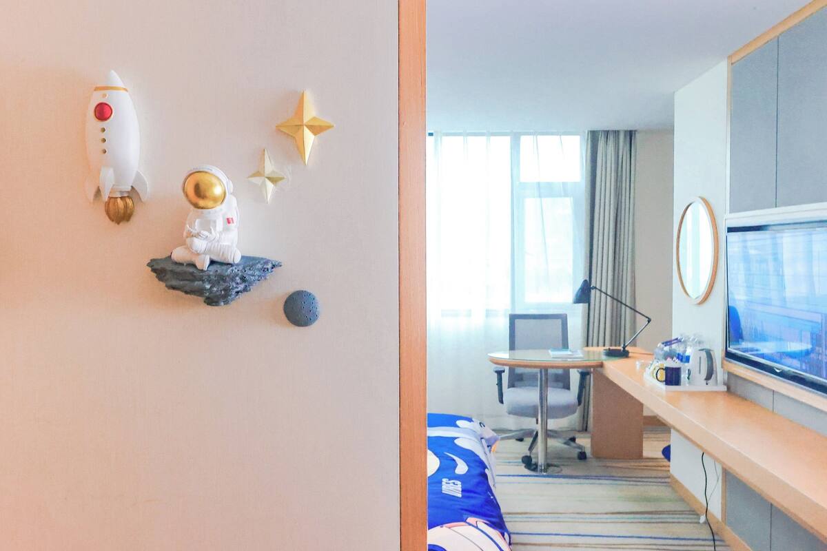 A hotel room with a bed, a desk, a television, and wall decorations including a rocket and astronaut figurine.