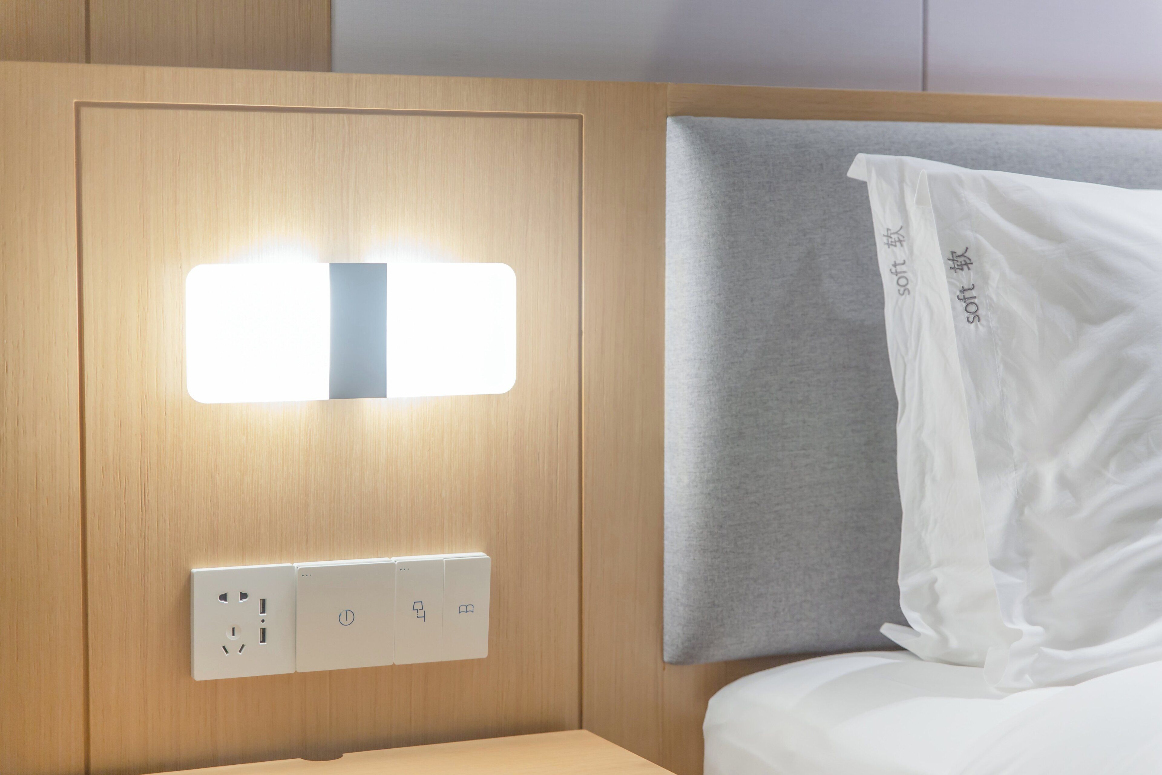 Ein Hotelzimmer mit einem hölzernen Kopfteil, einem Nachttisch mit Steckdose und einem weißen Kissen mit dem Aufdruck 'soft'.
