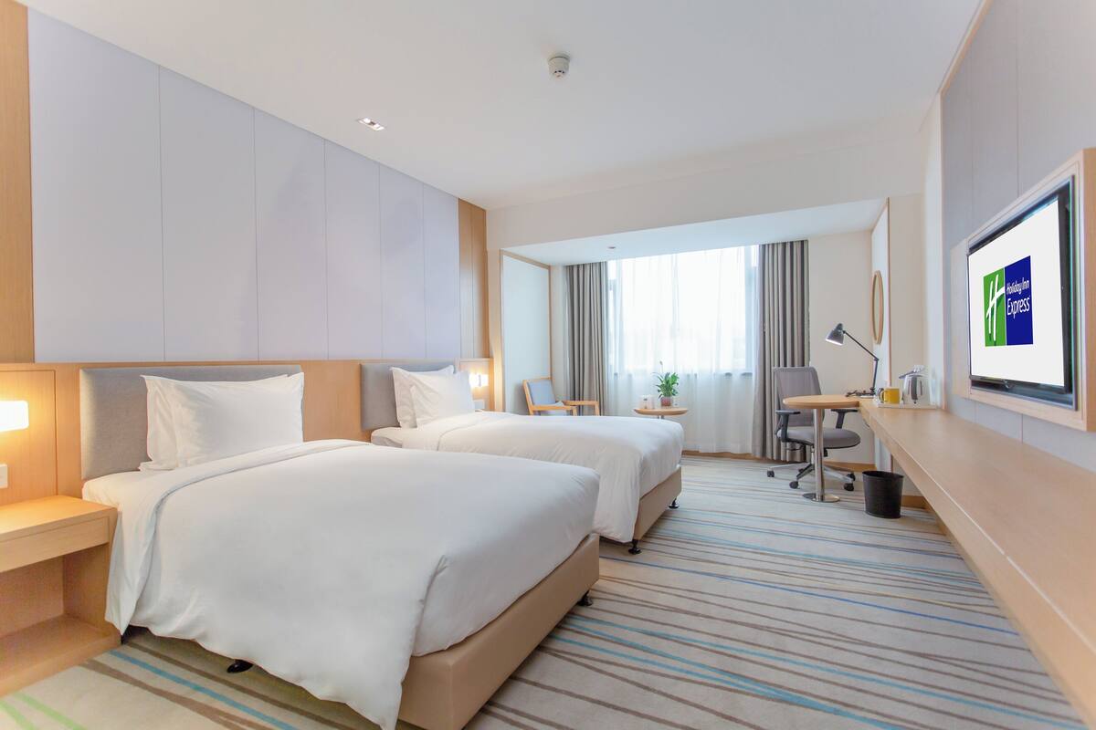 A hotel room with two beds, a desk, a chair, a TV, and a large window with curtains.