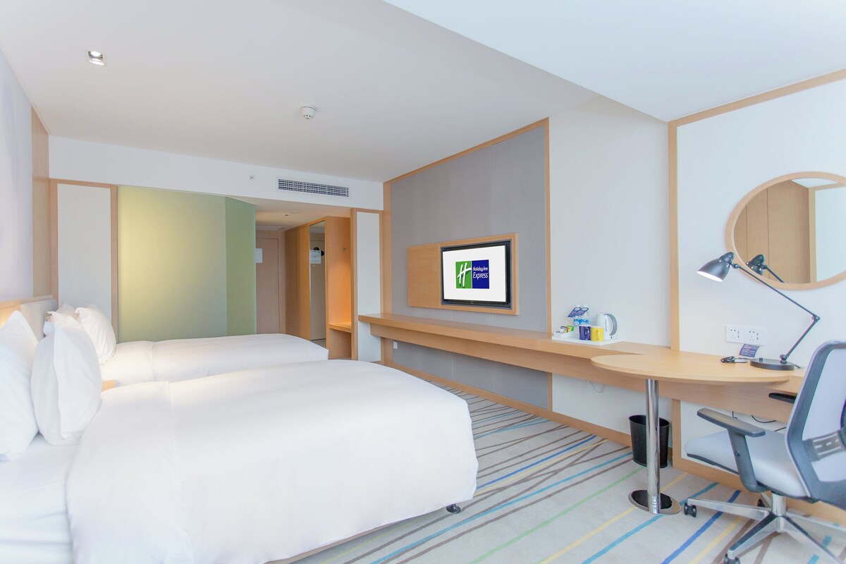 A hotel room with a bed, a desk, a chair, a desk lamp, a computer, and a television.