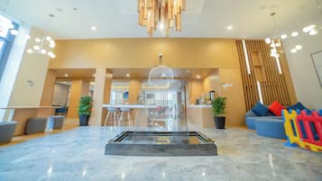 Een moderne lobby met een centrale glazen tafel, zithoekjes en een decoratieve plafondlamp.
