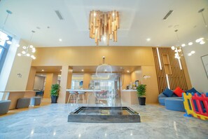 Een moderne lobby met een centrale glazen tafel, zithoekjes en een decoratieve plafondlamp.