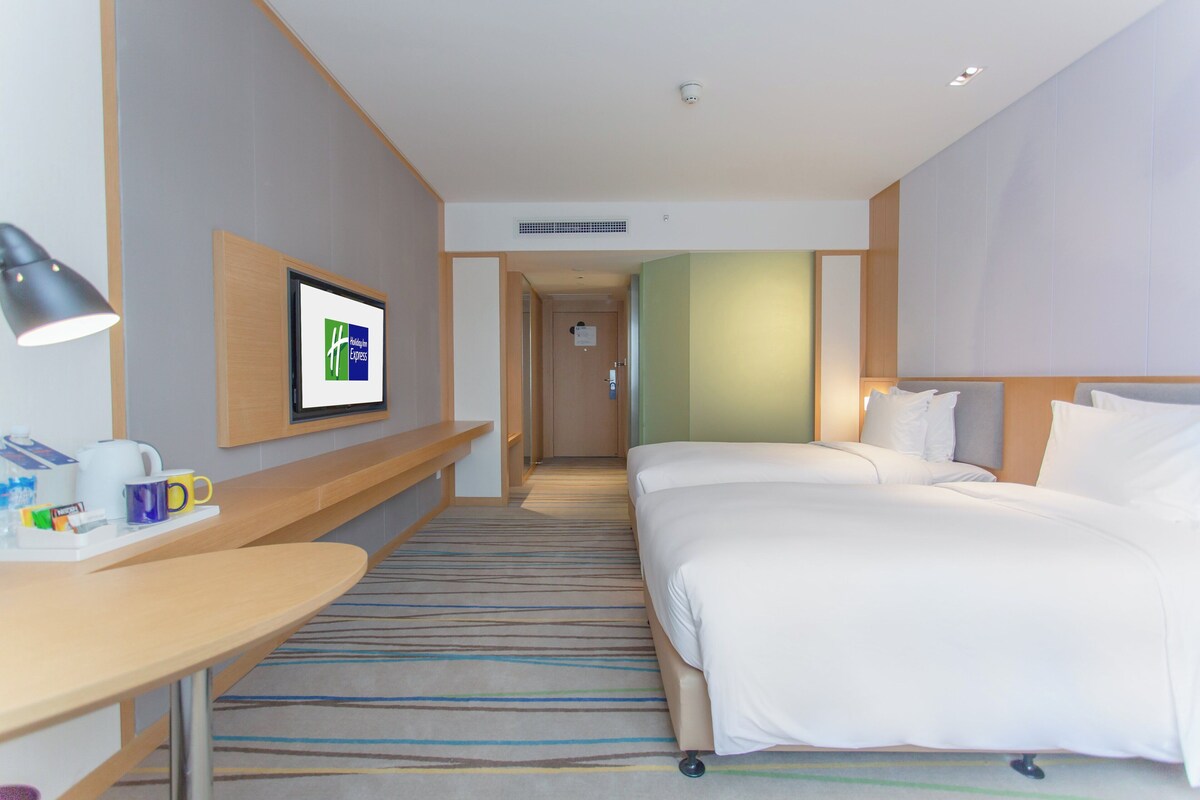 A hotel room with a bed, a desk with a lamp, a television, and a bathroom door in the background.