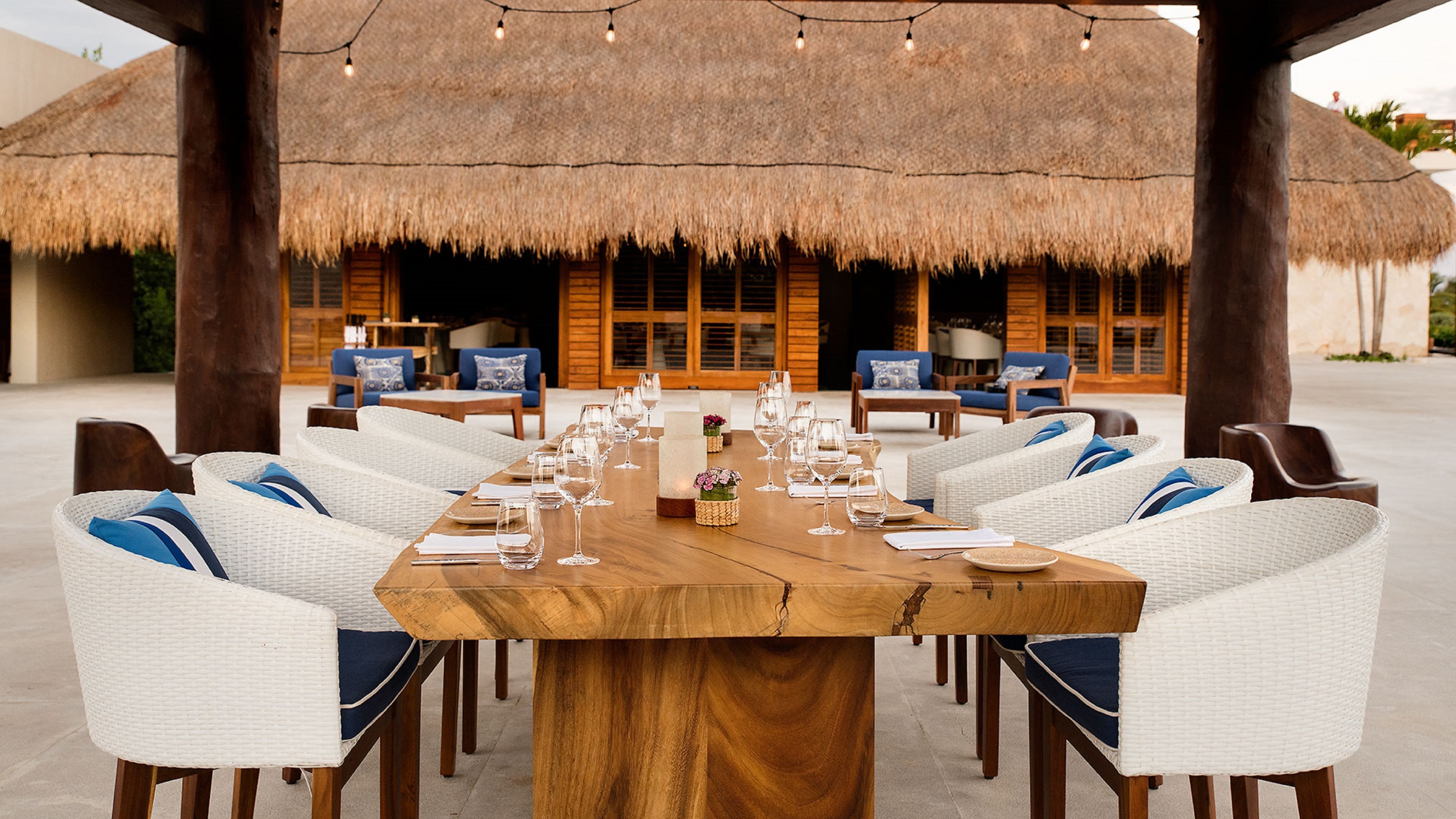A dining area with a large wooden table set for a meal, surrounded by white wicker chairs with blue cushions, under a thatched roof.