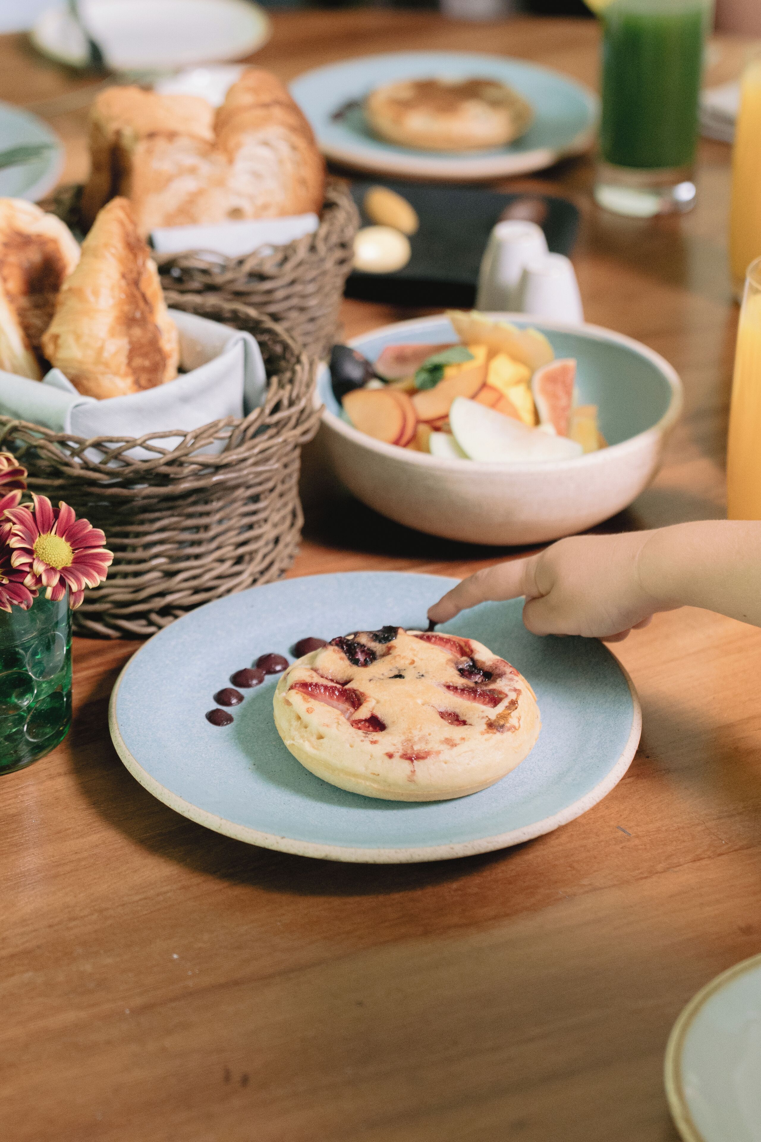 A breakfast setting with croissants, a fruit bowl, and a glass of green juice on a wooden table.
